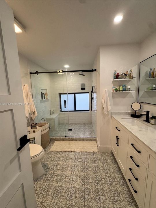 Custom cabinetry with dark hardware, stone-top vanity, walk-in glass-enclosed shower with matte black fixtures, patterned tile flooring, and integrated wall shelving