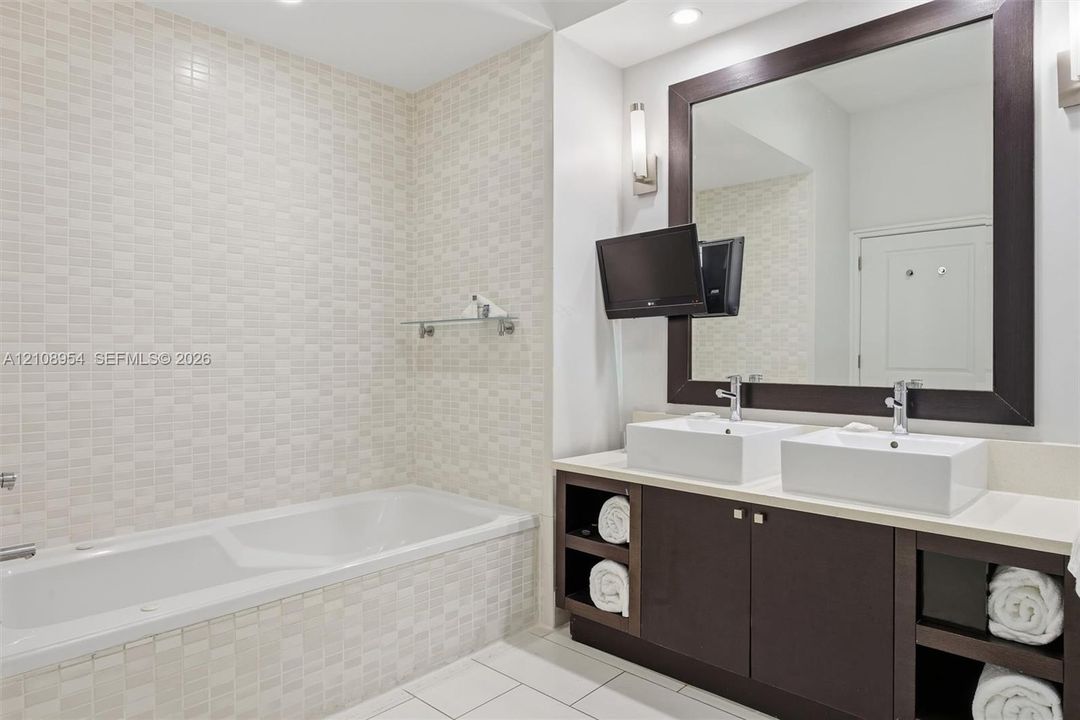 Dual vanity with rectangular vessel sinks, quartz-finish countertop, dark-toned cabinetry with open shelving, framed mirror, and tiled shower surround with built-in glass shelving