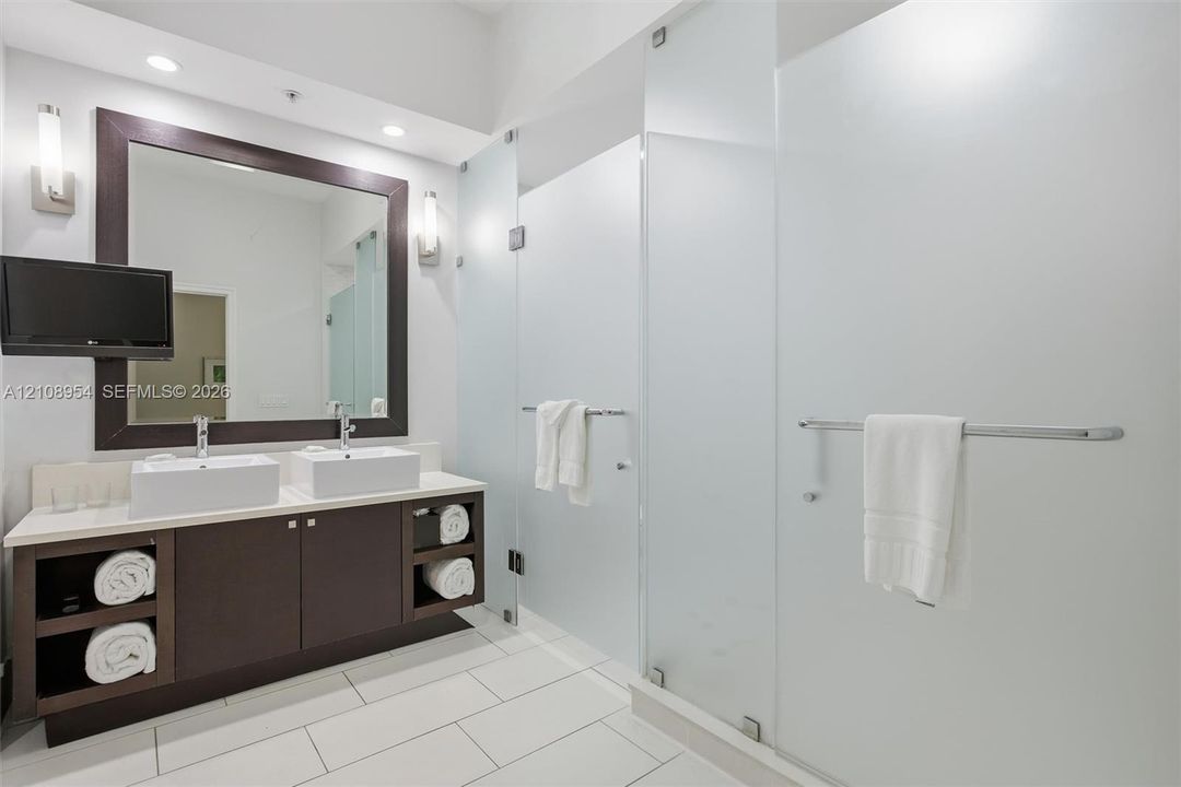 Dark-finish floating vanity with dual rectangular vessel sinks, light-toned countertop, framed mirror, and frosted glass shower enclosures with integrated metal hardware