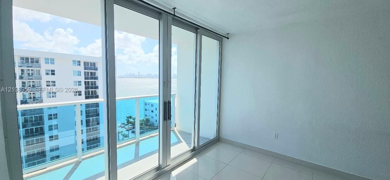 Floor-to-ceiling glass sliding doors, private balcony with glass railing, large-format tile flooring, and water backdrop
