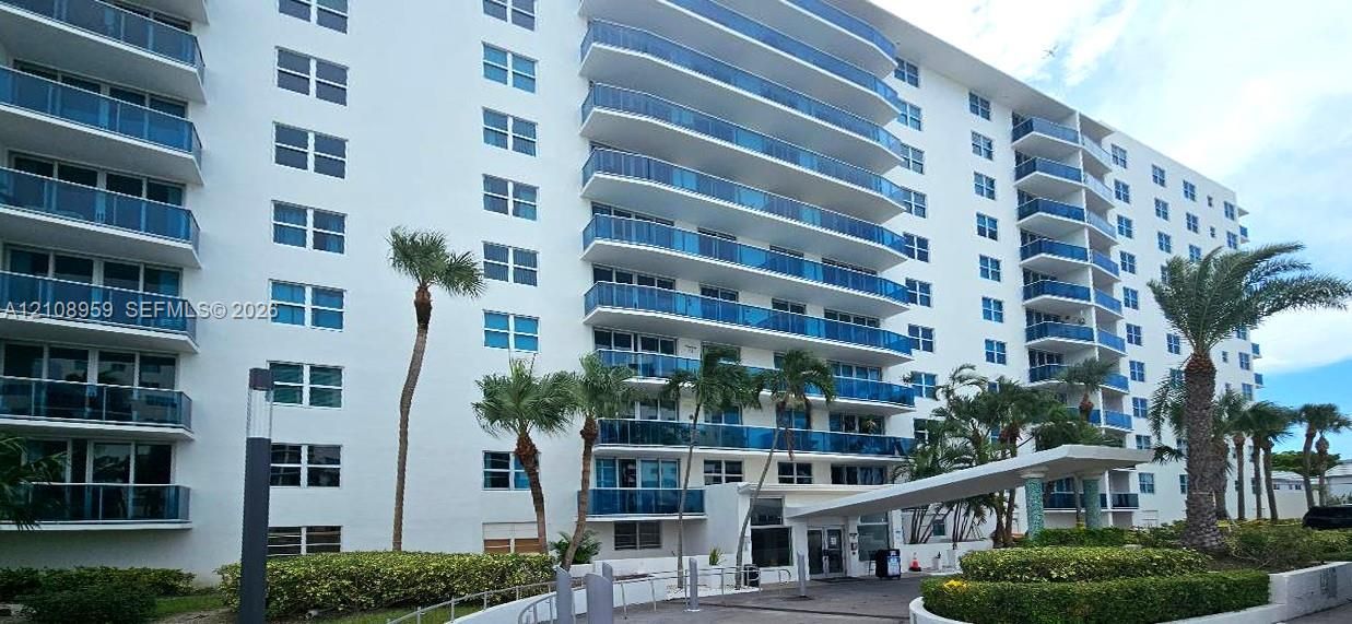 Multi-story residential building featuring private glass-railed balconies, a covered porte-cochère entry, and landscaped grounds with mature palm trees