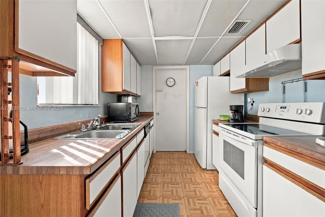 Kitchen featuring wood-trim cabinetry, laminate countertops, a dual-basin stainless steel sink, tile flooring, and integrated white appliances