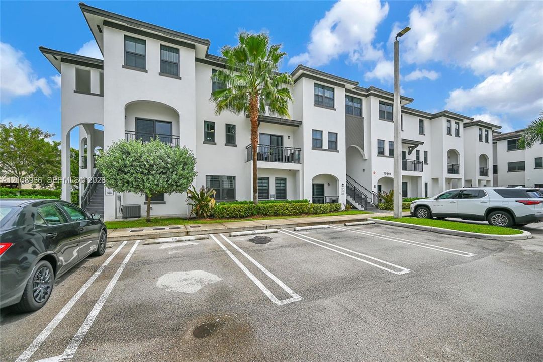 Multi-story stucco exterior featuring arched entryway detailing, private balconies with metal railings, designated surface parking, and manicured tropical landscaping