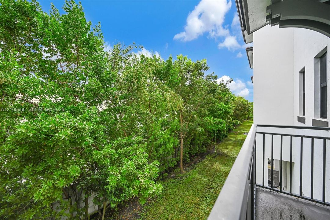 Elevated balcony featuring a metal railing, smooth stucco exterior, and concrete flooring, overlooking a green space with mature trees