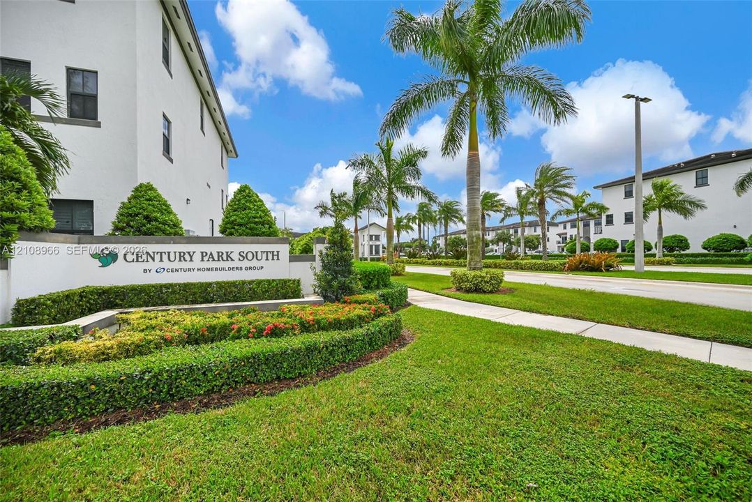 Community signage with stucco exterior wall, concrete sidewalk, mature palm trees, and manicured landscaping