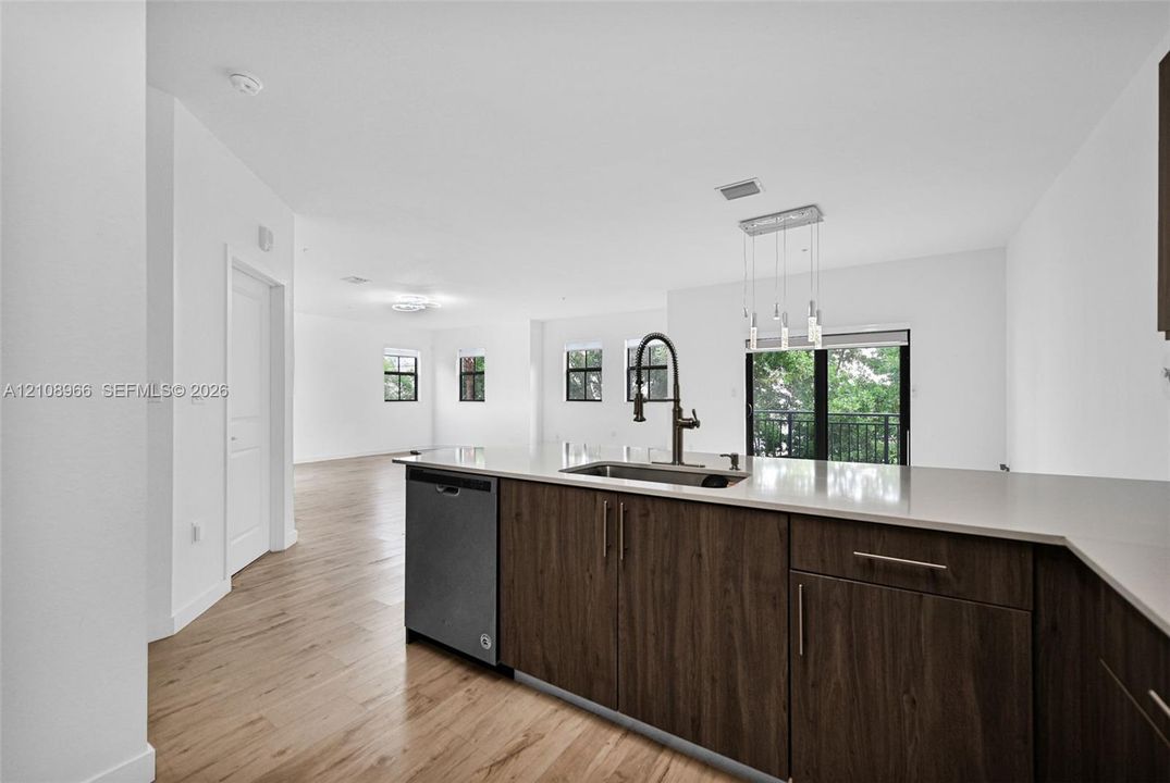 Kitchen island featuring wood-finish cabinetry, light-toned stone-style countertops, an undermount sink with a gooseneck faucet, integrated dishwasher, and hanging pendant lighting, complemented by wood-finish flooring