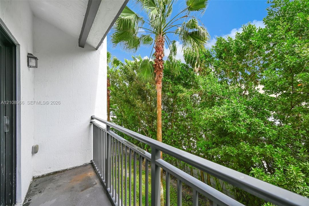 Covered balcony featuring a metal railing, exterior wall-mounted light fixture, and foliage