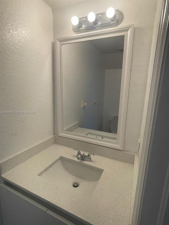 Vanity featuring a solid-surface countertop with an integrated basin, chrome-finish dual-handle faucet, framed wall mirror, and a three-light fixture