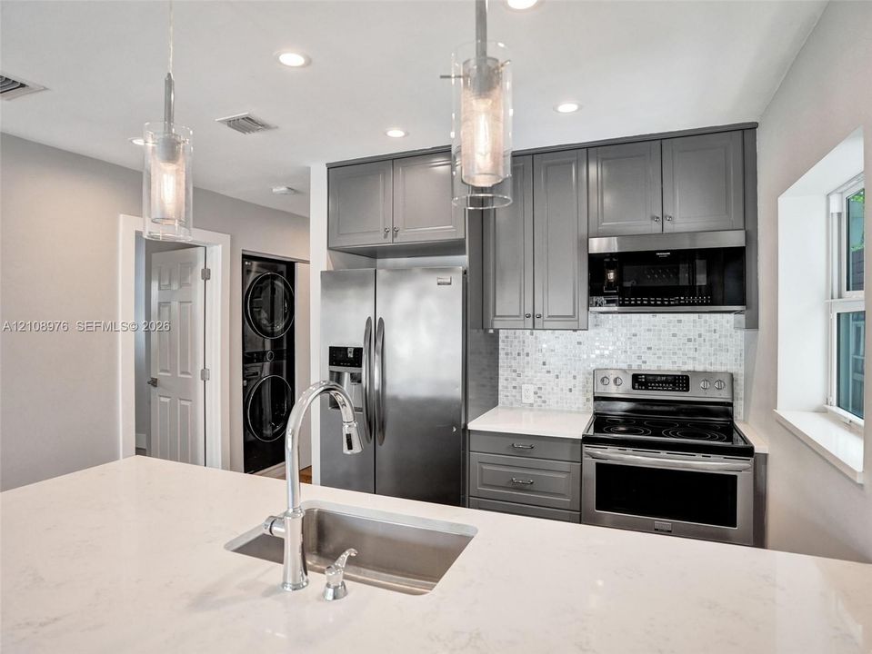 Kitchen featuring grey shaker-style cabinetry, white stone-finish countertops, stainless steel appliances, mosaic tile backsplash, and glass pendant lighting