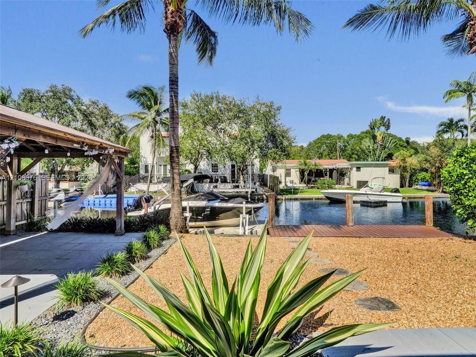 Waterfront property featuring a covered pergola, private wood-plank dock, gravel landscaping, and mature palm trees