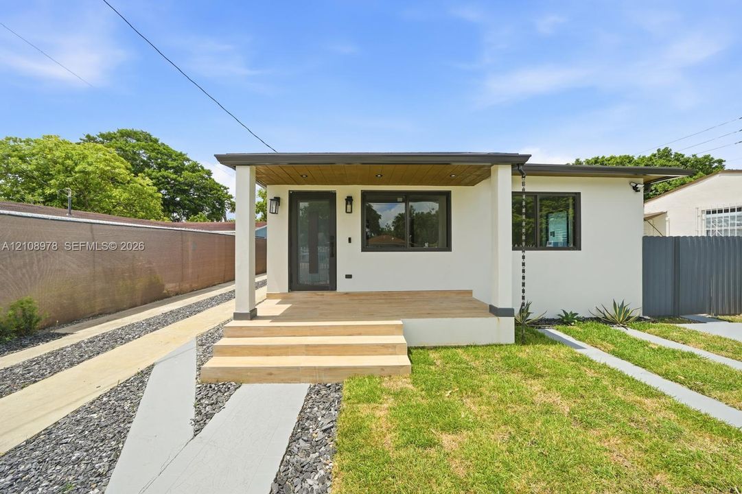Single-story exterior featuring a covered porch with wood-finish decking, recessed ceiling lighting, a contemporary glass-paneled entry door, and a metal rain chain