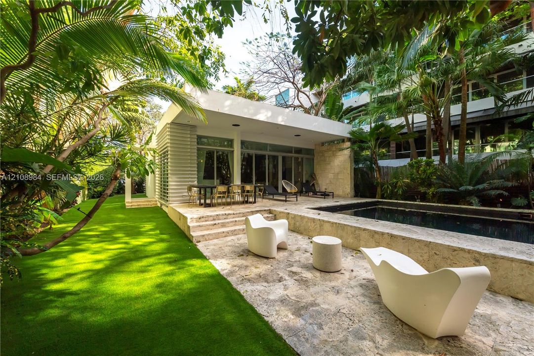 Outdoor space featuring a swimming pool, natural stone patio, covered terrace, and manicured lawn surrounded by mature palms and foliage