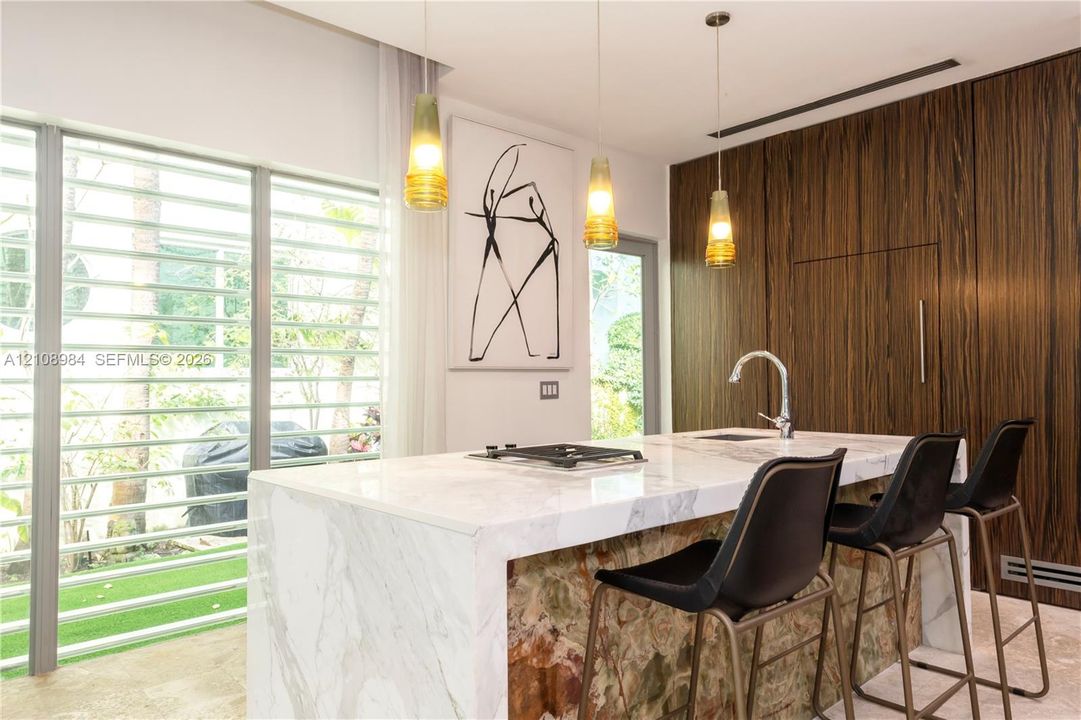 Kitchen island with stone countertop, waterfall edge, and built-in cooktop, paired with full-height wood-finish cabinetry and three pendant lights
