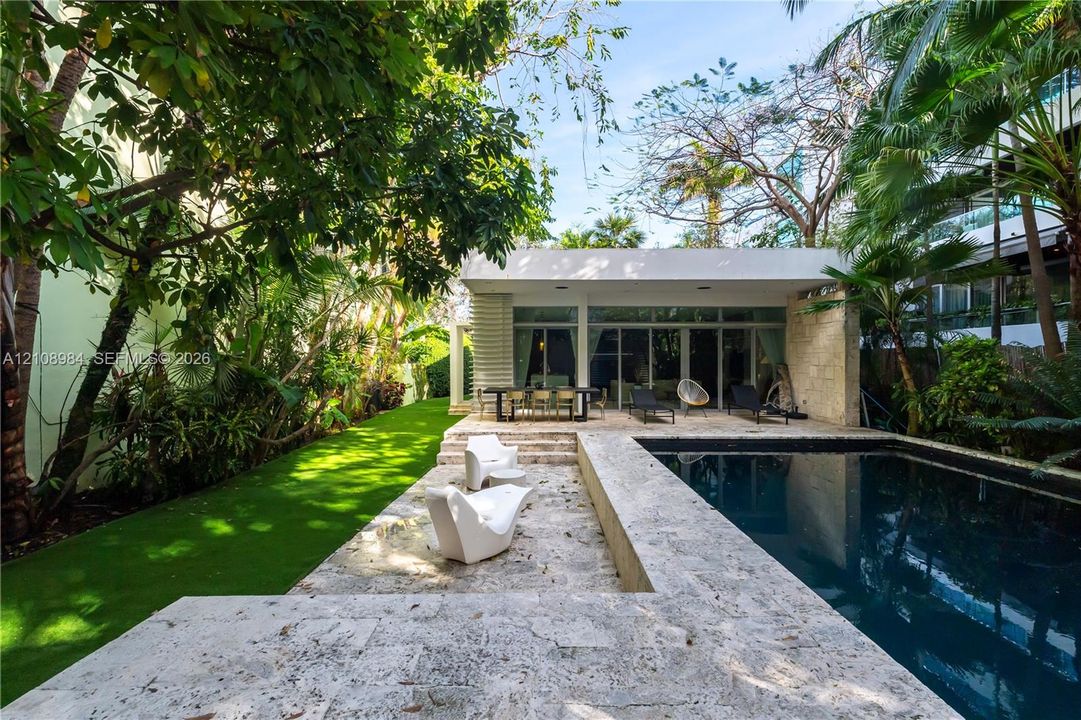 Outdoor living area featuring a custom swimming pool with stone decking, integrated landscape design, and a flat-roofed structure with floor-to-ceiling glass paneling