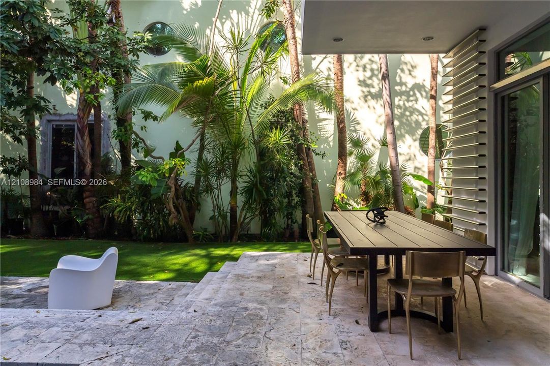Covered patio featuring stone tile flooring, structural louvered privacy screen, synthetic turf lawn, mature palms, and circular wall accents
