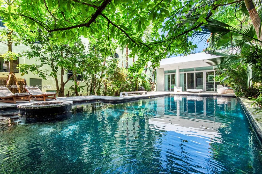 In-ground swimming pool featuring stone coping and a circular integrated spa, surrounded by mature trees, lush tropical landscaping, and expansive stone decking