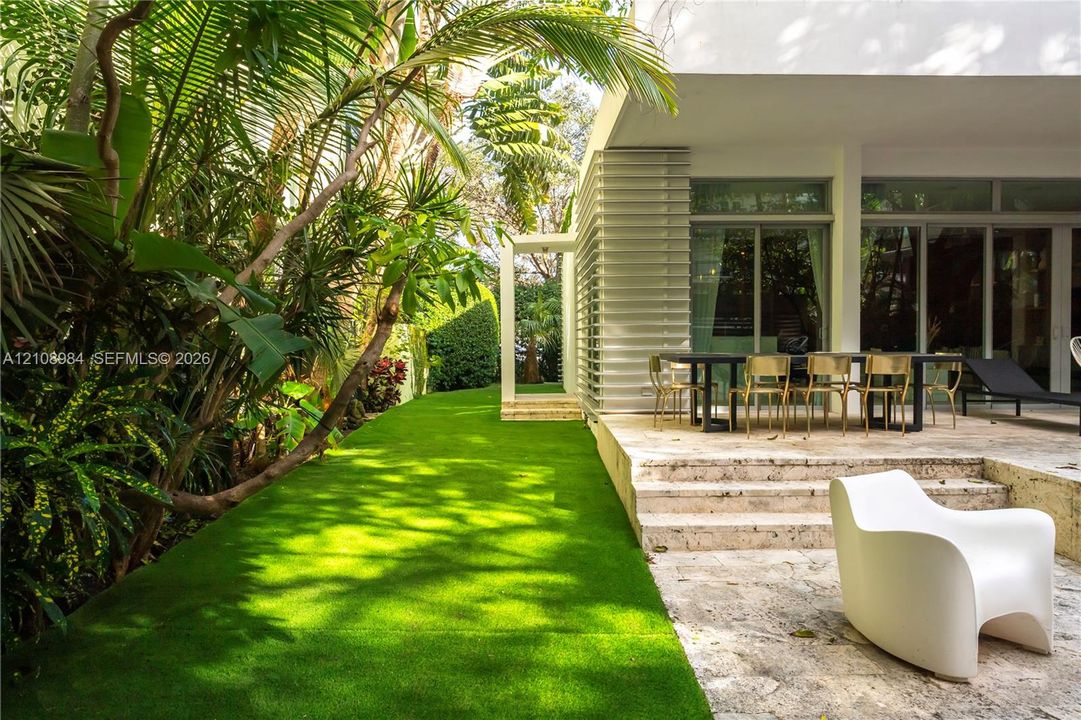 Natural stone paved patio featuring a built-in step, multi-panel sliding glass doors, horizontal exterior louvers, and a bordering artificial turf lawn with mature tropical landscaping