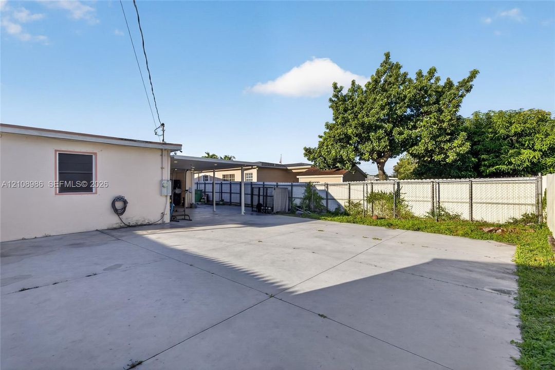 Expansive concrete paved exterior area featuring a covered carport, perimeter chain-link fencing, and established mature trees
