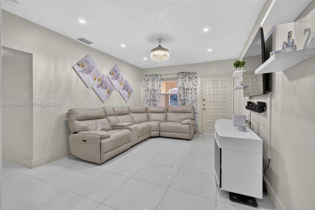 Living area featuring large-format tile flooring, recessed ceiling lighting, a decorative crystal-style chandelier, wall-mounted shelving, and a six-panel entry door