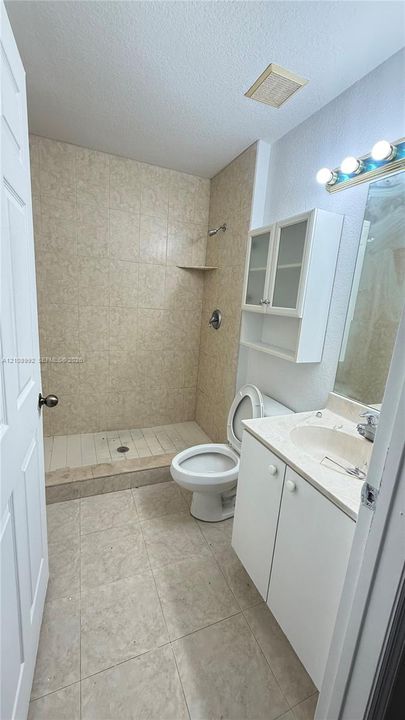 Full bathroom featuring a walk-in shower with tiled surround, stone-composite countertop, white cabinetry with integrated upper storage, and wall-mounted vanity lighting