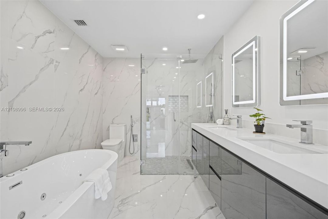 Full-wall veined stone-look tiling, jetted soaking tub, dual-sink vanity with high-gloss cabinetry, glass-enclosed walk-in shower with stone tile flooring, and integrated LED-lighted mirrors