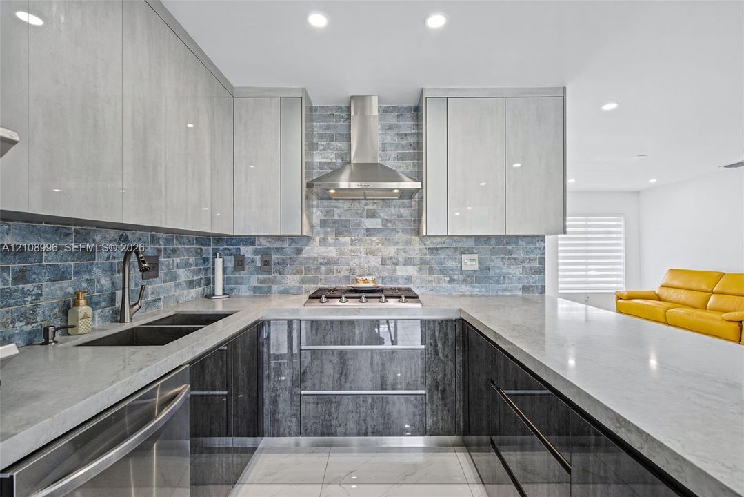 Kitchen featuring two-tone flat-panel cabinetry, stone-finish countertops, brick-style tile backsplash, stainless steel gas cooktop with chimney-style range hood, and integrated stainless steel sink with pull-down faucet