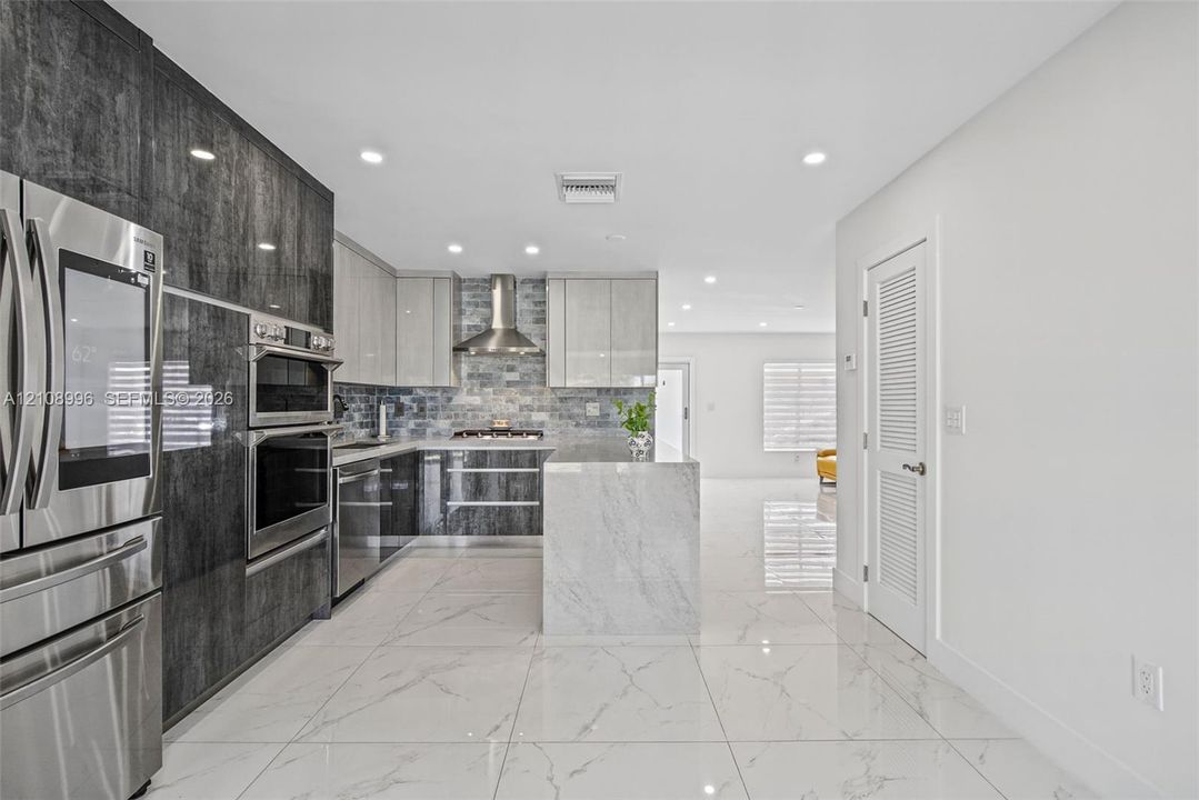 Kitchen featuring two-tone cabinetry with high-gloss finishes, a center island with stone-finish waterfall edges, stainless steel built-in appliances, a glass tile backsplash, and large-format porcelain tile flooring