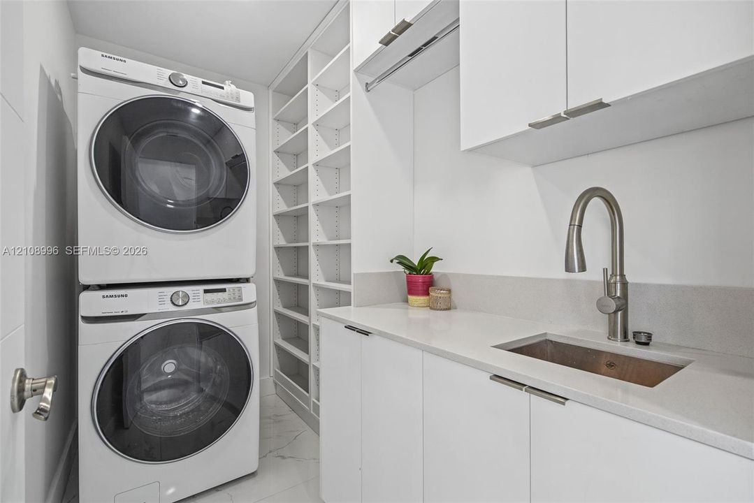 Laundry area featuring custom white cabinetry, stone-surface countertop with an integrated basin, high-arc pull-down faucet, floor-to-ceiling built-in shelving, and large-format tile flooring