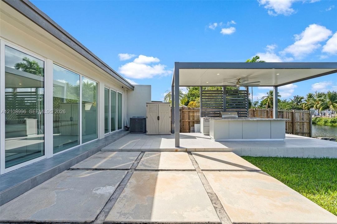 Outdoor patio featuring large-format concrete pavers, a covered pergola with recessed lighting and ceiling fan, a built-in outdoor kitchen with countertop, and floor-to-ceiling glass panels