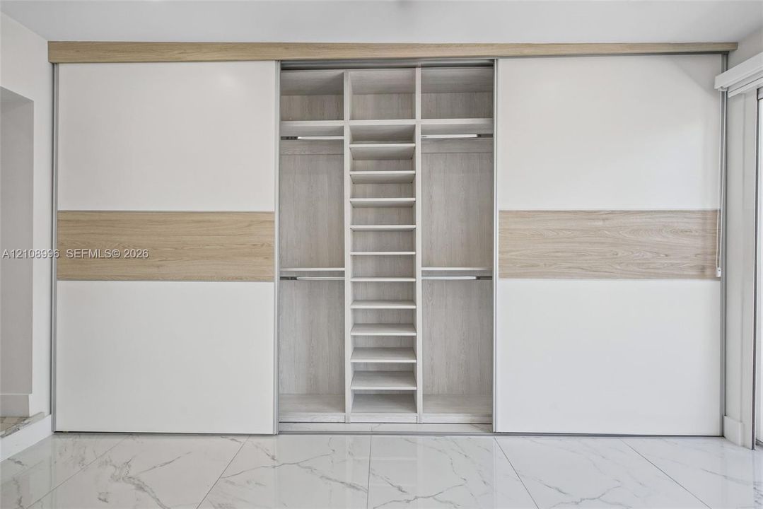 Built-in floor-to-ceiling wardrobe featuring sliding dual-tone panel doors, integrated shelving, hanging rods, and light wood-finish interior detailing, complemented by large-format polished tile flooring