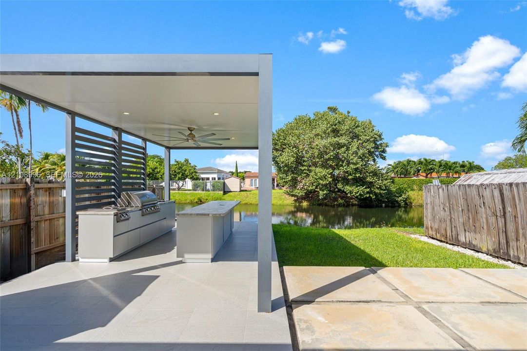 Outdoor kitchen featuring a built-in stainless steel grill, stone-topped island, modern pergola structure with recessed lighting and a ceiling fan, and large-format paved patio flooring