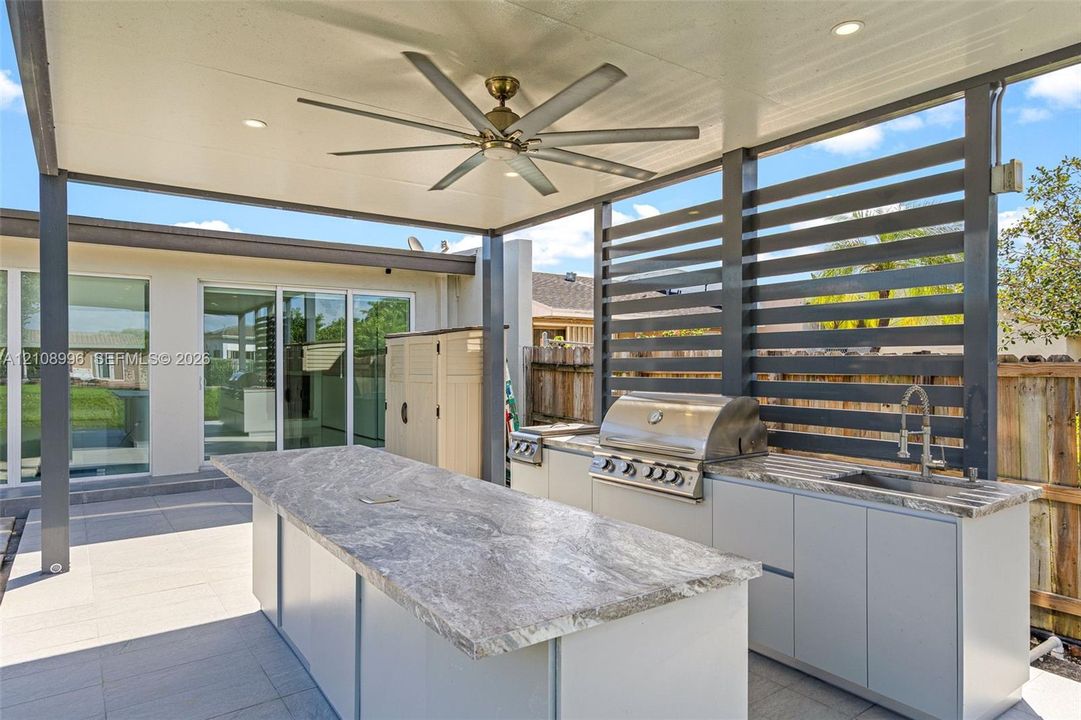 Covered outdoor kitchen featuring stone-top counters, a stainless steel built-in grill, a sink, and a multi-blade ceiling fan under a structured patio roof