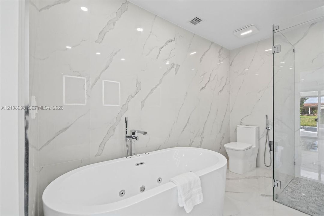 Freestanding jetted soaking tub with deck-mounted chrome faucet, floor-to-ceiling large-format stone-finish tile wall, frameless glass enclosed shower, and white dual-flush toilet