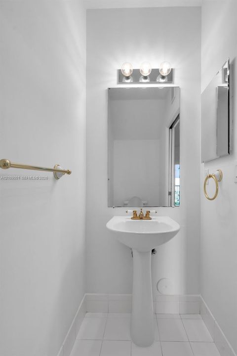 White pedestal sink with metallic gold-tone hardware, rectangular wall mirror, three-light vanity fixture, and matching gold-tone wall-mounted towel accessories