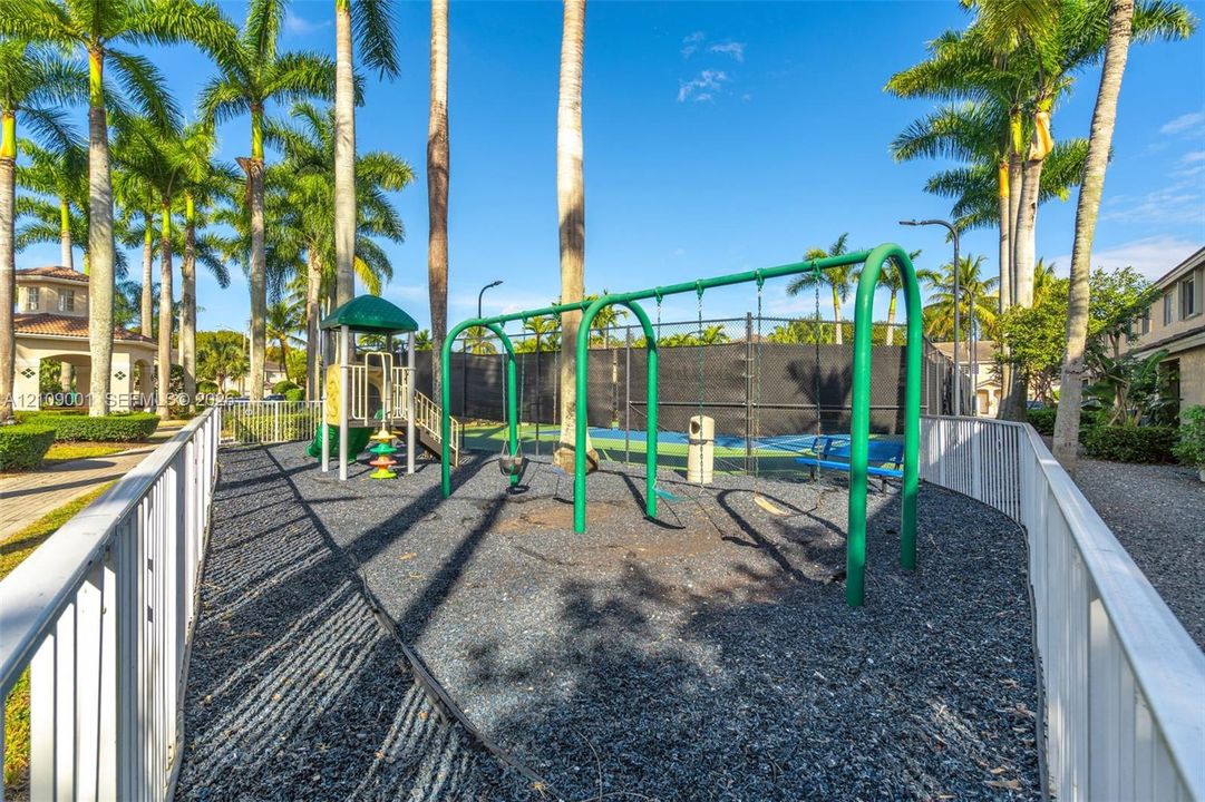 Outdoor recreational area featuring a playground with a swing set and slide, enclosed tennis court with perimeter fencing, and mature palm trees