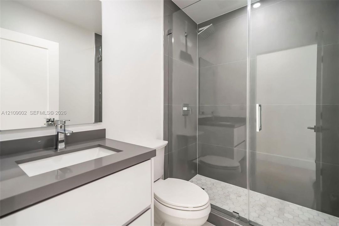 Modern vanity with rectangular sink and dark composite countertop, frameless glass shower enclosure, oversized wall tiling, and hexagonal floor tile in the shower