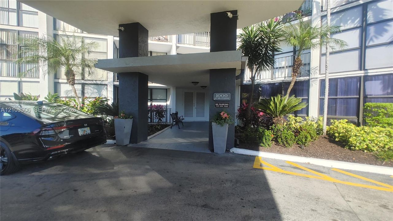 Covered building entrance featuring a concrete porte-cochère, decorative structural pillars, and curated landscape planters