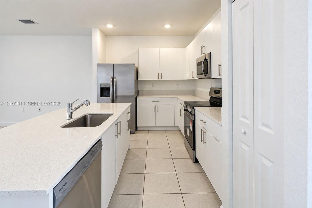 Kitchen featuring white shaker-style cabinetry, solid-surface countertops, a center island with an integrated stainless steel sink, stainless steel appliances, and tile flooring