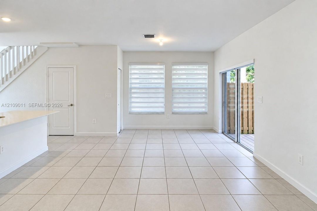 Open-concept interior featuring neutral ceramic tile flooring, sliding glass patio doors, dual windows with integrated shades, and a staircase with a white railing