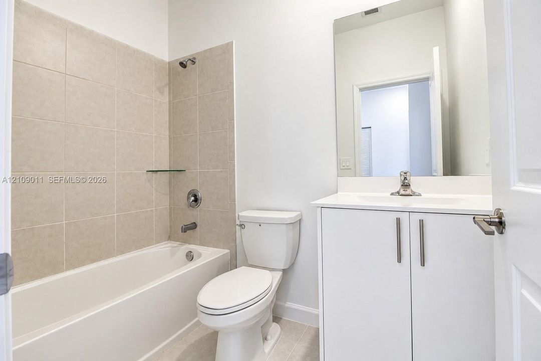 Bathroom featuring a white vanity cabinet with an integrated sink, a rectangular wall-mounted mirror, a tub-shower combination with neutral tile surround, and a glass shelving unit