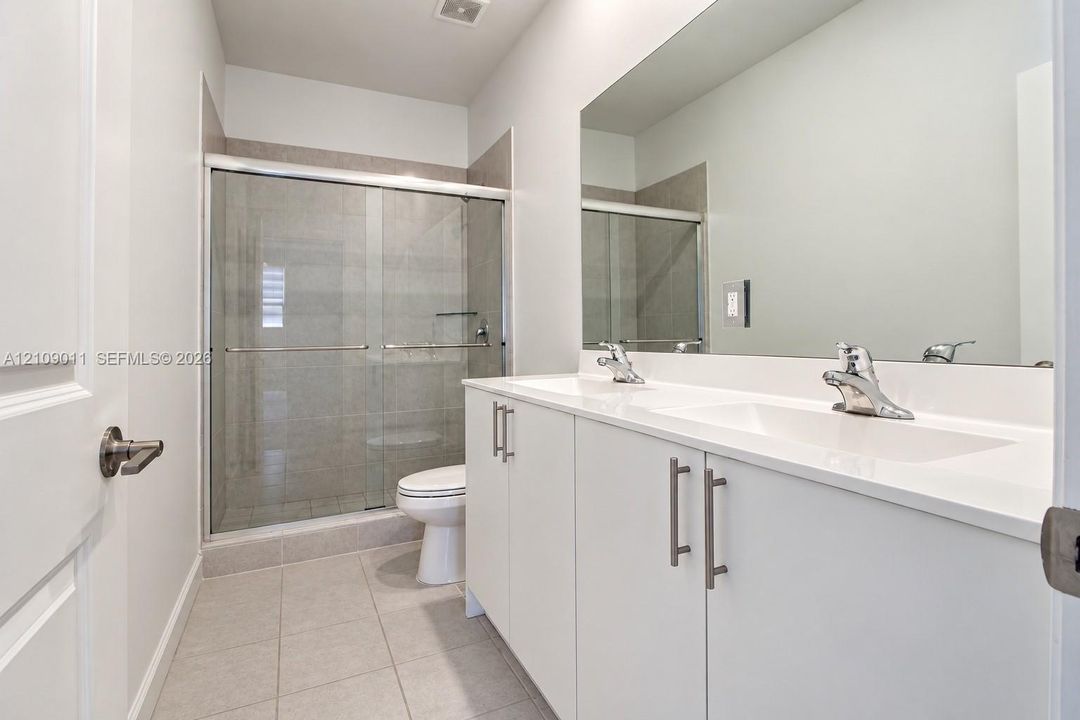 Double-sink vanity with white cabinetry, solid surface countertop, glass-enclosed shower, and tile flooring