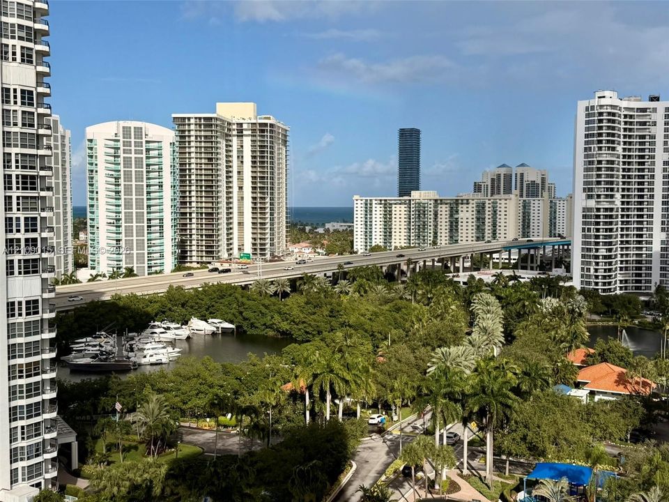 Panoramic cityscape featuring high-rise architecture, a coastal waterway with boat docks, lush palm trees, and an ocean horizon