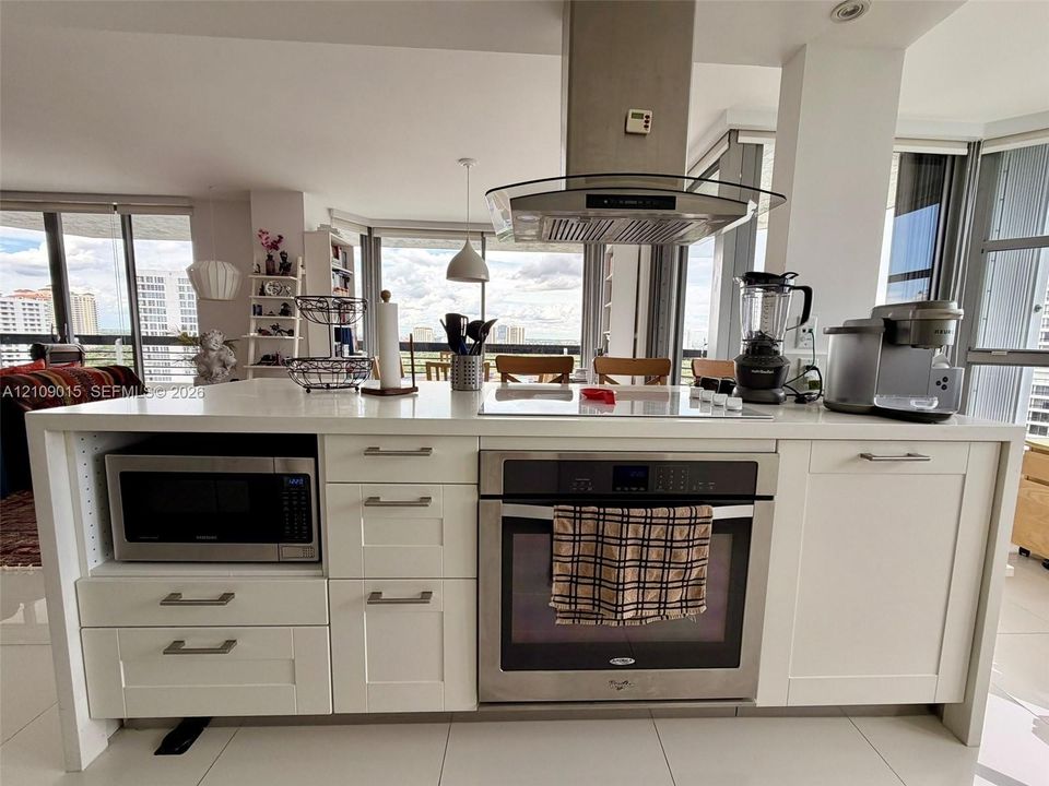 Kitchen island featuring white cabinetry, a light-toned solid surface countertop, stainless steel oven, integrated microwave, and an overhead stainless steel range hood