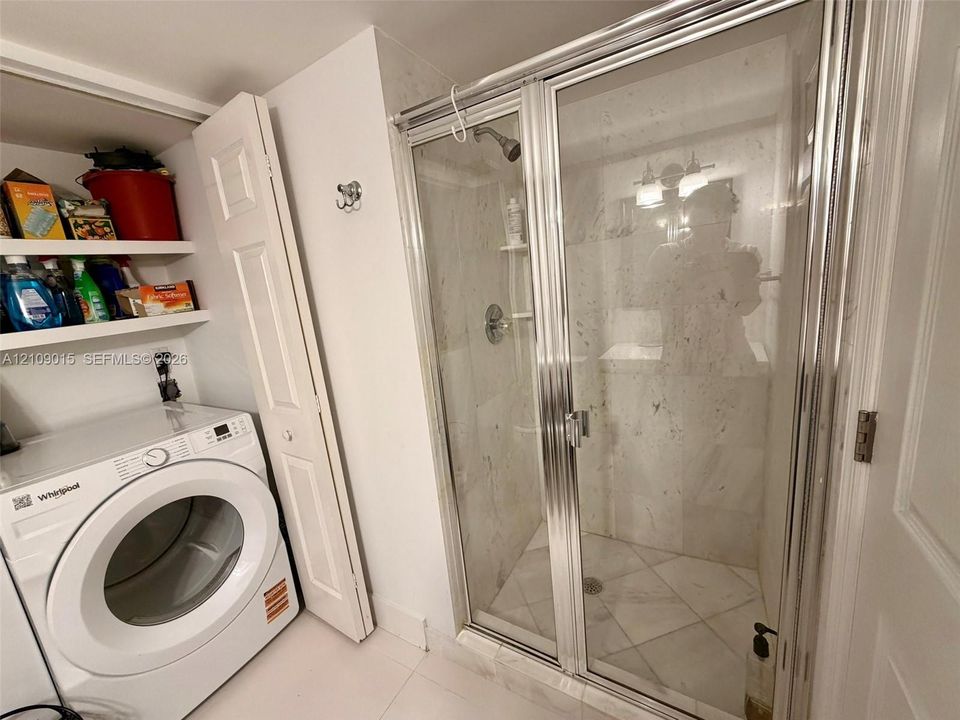 Walk-in shower enclosure with glass doors, stone-finish wall tiling, built-in shelving, chrome-finish plumbing fixtures, and integrated laundry area with overhead storage shelving