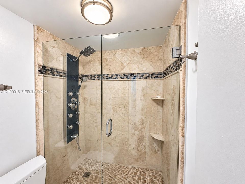 Walk-in shower featuring natural stone wall tile, decorative mosaic glass accent border, frameless glass door, integrated multi-head shower panel, and built-in corner shelving