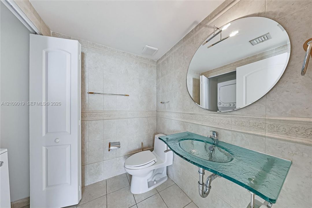 Bathroom featuring full-height stone-finish wall tile with decorative inlay, custom glass vanity top with integrated sink, oval wall-mounted mirror, and floor-to-ceiling storage cabinetry