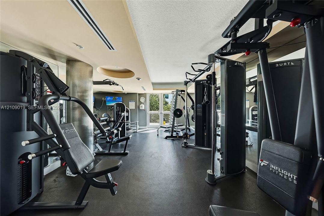 Fitness room featuring high-durability rubber flooring, metallic structural columns, recessed ceiling lighting, and a glass-paneled exterior door