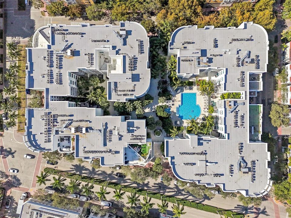 Aerial perspective of a multi-story residential complex featuring a central outdoor swimming pool, landscaped courtyards with palm trees, paved walkways, and extensive surrounding greenery