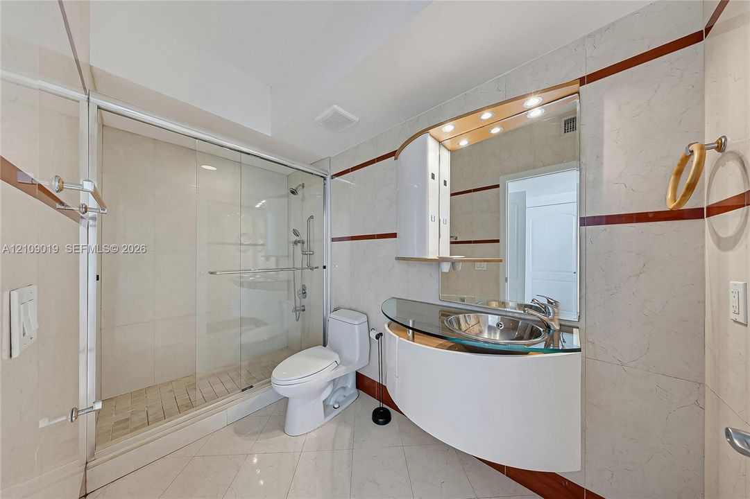 Curved wall-mounted vanity with a glass countertop, integrated vessel sink, glass-enclosed shower with tiled walls, and large-format tile flooring with decorative inlay