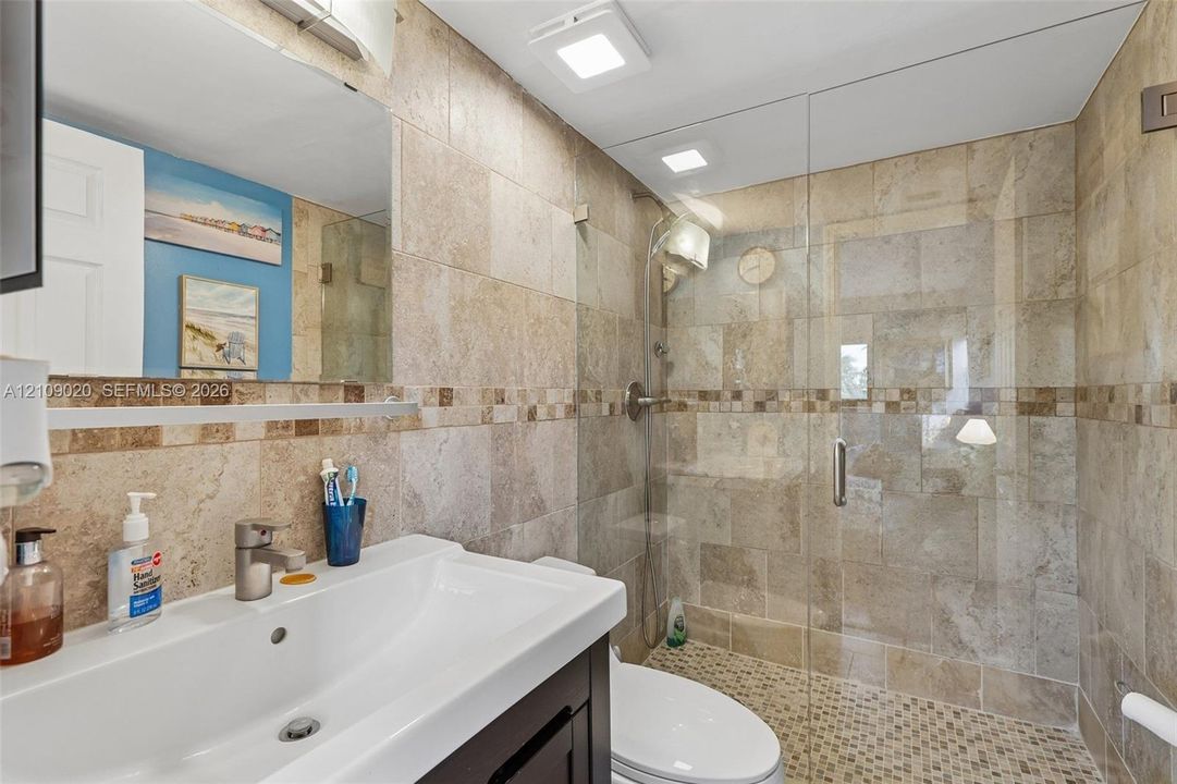 Bathroom featuring large-format natural stone wall tiling with mosaic accent banding, a glass-enclosed shower with pebble tile flooring, and a dark-finish vanity with an integrated basin